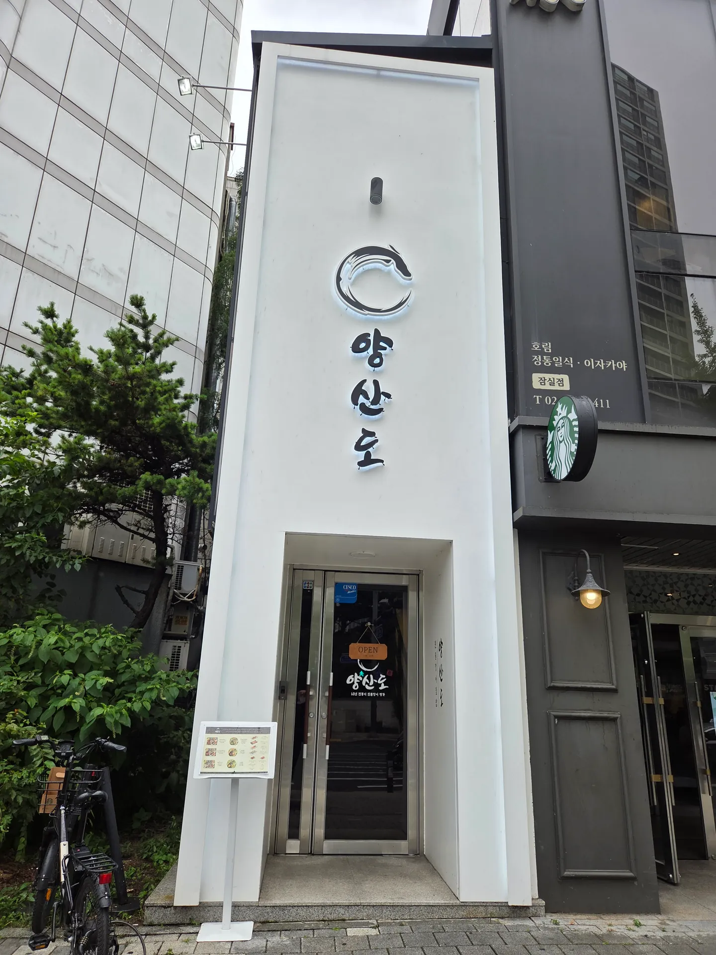 상단 돌출 표식과 밝은 외벽 및 하부 유리 출입구가 이어진 매장 전면